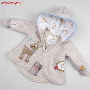 May include: A beige hooded jacket with a front zipper, featuring a brown deer appliqué on the chest and a light blue butterfly. The hood lining has a repeating deer pattern. A circular patch on the inside reads Emma. A small tag is attached to the zipper.