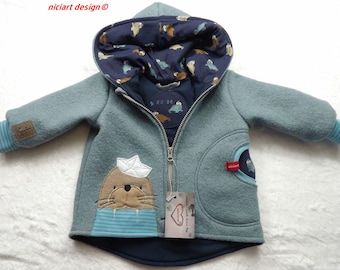 Peut inclure: Une veste à capuche bleu clair avec une fermeture éclair, ornée d'un appliqué de morse beige portant un chapeau en bateau de papier blanc. La doublure de la capuche est bleu marine avec un motif de morse. Les poignets sont rayés et une étiquette pend.