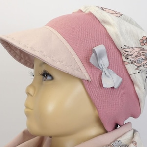 Op de afbeelding: Een roze en beige babyhoed met een visor en een grijze strik. De hoed heeft een bloemenpatroon op de achterkant.