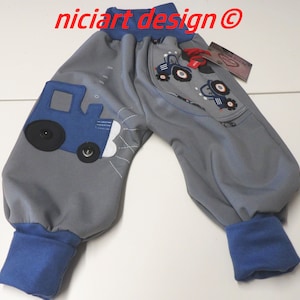 Può includere: Pantaloni della tuta grigi con polsini blu e un'applicazione di trattore blu. Il trattore ha ruote nere e fari bianchi. I pantaloni della tuta hanno un piccolo cartellino rosso con il testo "niciart design@" sul lato destro.