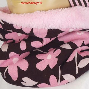 Op de afbeelding: Een nekwarmer met een donkerbruine basis en een herhalend patroon van lichtroze bloemen en stengels. De binnenzijde is gevoerd met zacht, lichtroze imitatiebont. De tekst "niciart design ©" is in het rood bovenaan in het midden gedrukt.