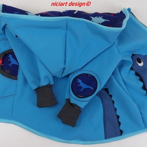 Może przedstawiać: Niebieska kurtka z kapturem i wzorem dinozaura. Kurtka ma zamek błyskawiczny i aplikację dinozaura z przodu. Kaptur jest podszyty niebieskim i białym materiałem z nadrukiem dinozaurów. Tekst "nicicart design®" jest widoczny na kapturze.