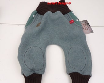 May include: Teal blue fleece baby pants with brown ribbed cuffs and green fabric panels. The pants have knee pads and a white button on each side. The pants have a label that says "nicicart design ©".
