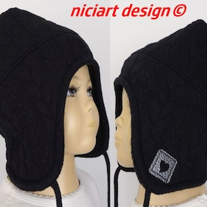 Può includere: Cappello invernale in pile nero con una toppa a forma di cuore e un pon pon in cima. Il cappello ha una patta che può essere tirata verso il basso per coprire le orecchie e il collo.