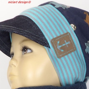 Op de afbeelding: Een blauwe en turquoise gestreepte beanie met een denim rand en een bruine patch met een ankerontwerp. De beanie heeft een patroon van cartoondieren op de achterkant.