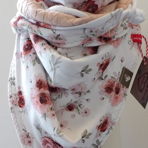 Op de afbeelding: Een witte sjaal met een bloemenpatroon met roze en rode rozen. De sjaal is gevouwen en heeft een roze laag eronder. De sjaal heeft een gehaakt bloemendetail en een label met de tekst "niciart design@" en "nicia".