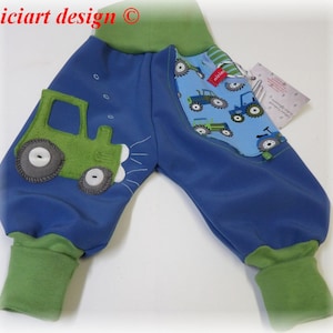 Può includere: Pantaloni per neonati blu con polsini verdi e un'applicazione di trattore verde sulla gamba sinistra. La gamba destra ha una tasca blu con una stampa di trattore.