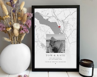 Puede incluir: Una impresión enmarcada de un mapa en blanco y negro con una foto de una pareja. El mapa resalta una ubicación con un corazón rojo. El texto en la parte inferior dice "TOM & KATE" y "YOU'RE MY GREATEST ADVENTURE". Un jarrón blanco con flores secas y una vela también están en la imagen.