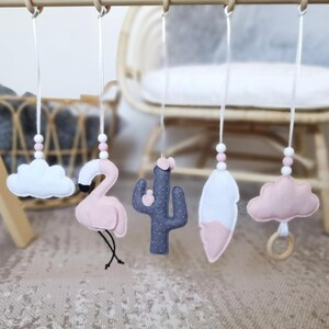 May include: A baby mobile with felt decorations: a white cloud, a pink flamingo, a grey cactus, a white and pink feather, and a pink cloud with a wooden ring. The shapes hang from a wooden bar with white string and pink and white beads.