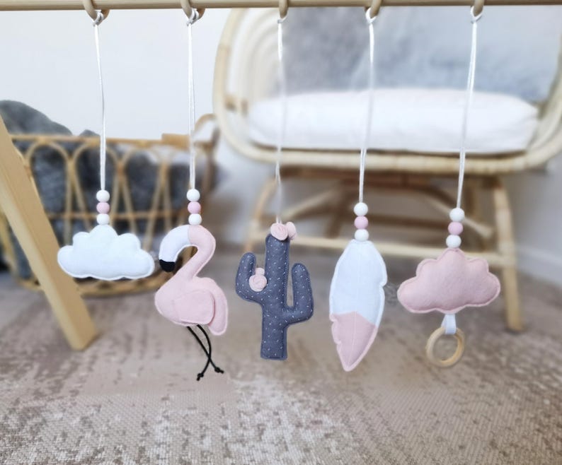 May include: A baby mobile with felt decorations: a white cloud, a pink flamingo, a grey cactus, a white and pink feather, and a pink cloud with a wooden ring. The shapes hang from a wooden bar with white string and pink and white beads.