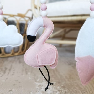 May include: A baby mobile with felt decorations, including a pink flamingo with a black beak, a white cloud, and a feather. The mobile has pink and white wooden beads and black strings.