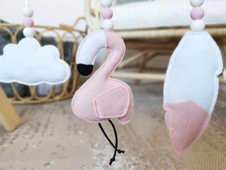 May include: A baby mobile with felt decorations, including a pink flamingo with a black beak, a white cloud, and a feather. The mobile has pink and white wooden beads and black strings.