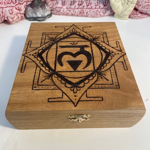 Puede incluir: Caja de madera marrón claro con un diseño de mandala negro en la tapa. La caja tiene un cierre dorado. El diseño incluye un símbolo central y patrones detallados, posiblemente para fines espirituales o decorativos.