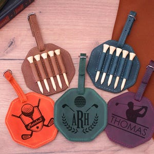 Puede incluir: Cinco portamarcadores de bolas de golf y porta-tees de cuero con nombres personalizados. Los portamarcadores son de varios colores, incluyendo naranja, verde, morado y marrón. Cada portamarcadores tiene un diseño diferente, incluyendo un palo de golf, una bola de golf y un golfista.