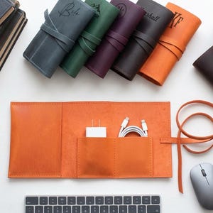 May include: A collection of leather roll-up organizers in various colors including blue, green, purple, brown, and orange. One orange organizer is laid flat, featuring two interior pockets holding a white power adapter and white charging cables.