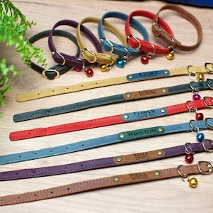 K&ouml;nnte beinhalten: Neun bunte Lederkatzenhalsb&auml;nder mit personalisierten Namensschildern und einem Gl&ouml;ckchen-Anh&auml;nger. Die Halsb&auml;nder sind in verschiedenen Farben, darunter Braun, Rot, Blau, Gr&uuml;n und Lila. Die Namensschilder sind mit den Namen "Cory", "Jessie", "Copper", "Winston", "Kelly", "Polly" und anderen Namen graviert.