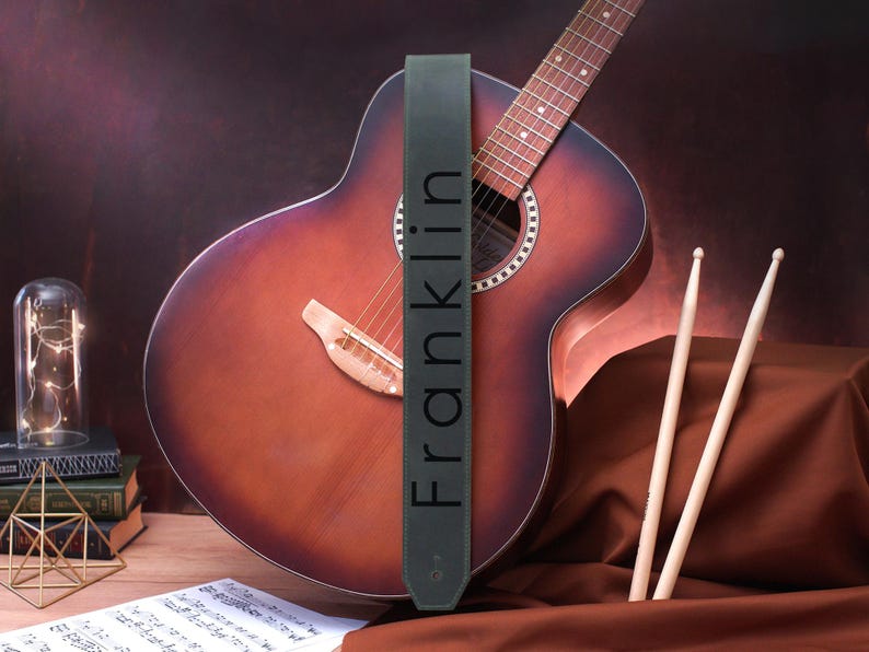 May include: A dark green leather guitar strap with the word "Franklin" printed in white on it. The strap is hanging on a brown acoustic guitar.