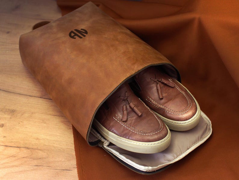 May include: A brown leather shoe bag with a zipper, holding a pair of brown leather shoes with white soles. The bag has a handle and the initials "AN" embossed on the front.