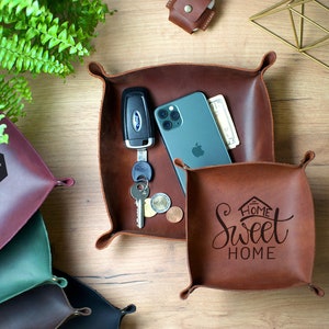 May include: Two brown leather catchall trays with different designs. One tray has the text "Home Sweet Home" and the other has the text "Dochel" and a house with a star and moon above it. The trays are on a wooden surface with a gold geometric ornament.