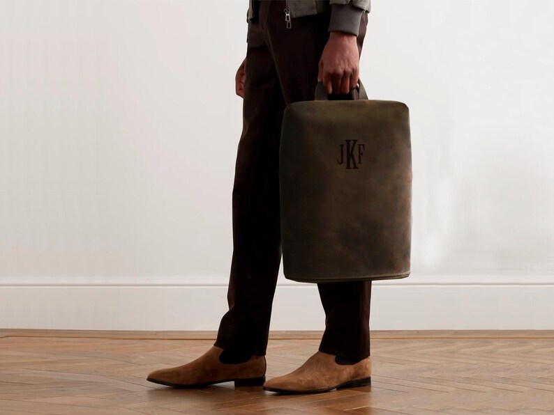 May include: A dark olive green leather tote bag with a handle, featuring the initials "JKF" in a darker shade. The bag is held by a person wearing brown pants and suede shoes.