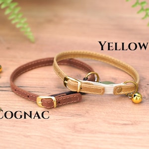 May include: Two cat collars, one brown and one yellow, with a bell on the yellow collar. The collars are on a wooden surface. The brown collar is labeled "COGNAC" and the yellow collar is labeled "YELLOW".