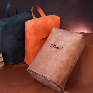 May include: Three leather shoe bags in black, orange, and brown, each with a handle and a name embroidered on the front. The brown bag has a zipper. The bags are on a brown surface.