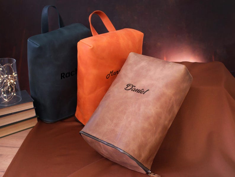 May include: Three leather shoe bags in black, orange, and brown, each with a handle and a name embroidered on the front. The brown bag has a zipper. The bags are on a brown surface.