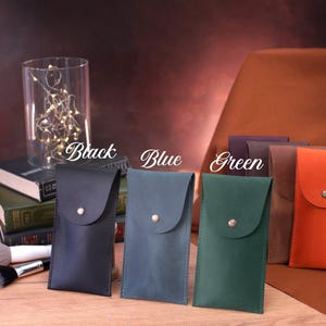 May include: Five leather phone cases in different colors: black, blue, green, brown, and orange. Each case has a flap with a button closure.