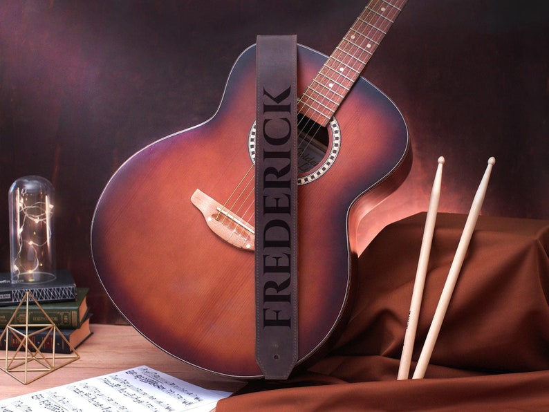 May include: A brown leather guitar strap with the name "FREDERICK" embossed in white lettering. The strap is attached to an acoustic guitar with a brown wood body and a natural wood fretboard.