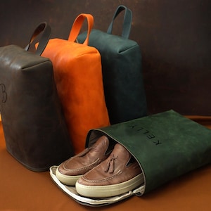 May include: Leather shoe bags in brown, orange, and teal, with matching handles. One bag is open, revealing brown loafers. The open bag has the name "KELLY" printed on it. A brown bag has the letter "B" on it.