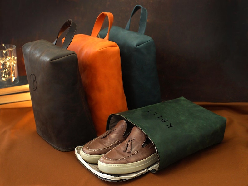 May include: Leather shoe bags in brown, orange, and teal, with matching handles. One bag is open, revealing brown loafers. The open bag has the name "KELLY" printed on it. A brown bag has the letter "B" on it.