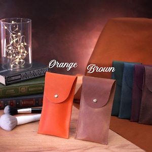 May include: Five leather phone cases in different colors, including orange, brown, teal, purple, and dark brown. The cases have a flap with a button closure. The cases are arranged on a wooden surface with a stack of books and a glass jar with lights behind them.