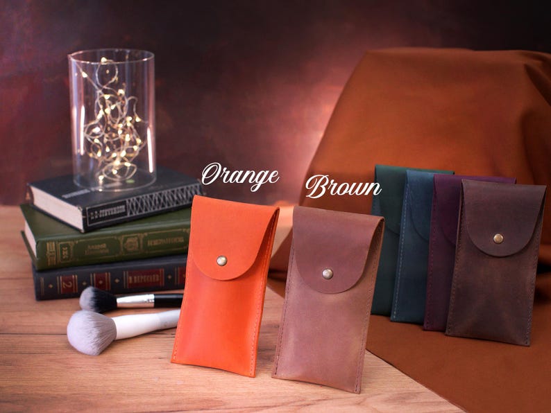 May include: Five leather phone cases in different colors, including orange, brown, teal, purple, and dark brown. The cases have a flap with a button closure. The cases are arranged on a wooden surface with a stack of books and a glass jar with lights behind them.