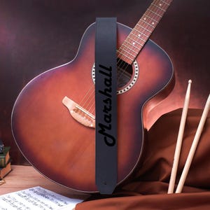May include: A black leather guitar strap with the word "Marshall" printed in white lettering. The strap is attached to a brown acoustic guitar.