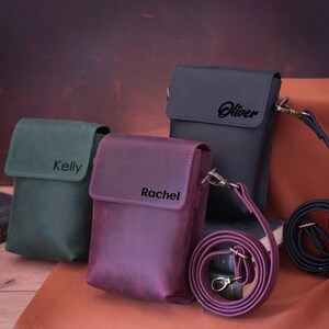 May include: Three leather crossbody bags in green, purple, and dark gray. Each bag has a flap closure and a name printed on the front: "Kelly", "Rachel", and "Oliver". The bags have adjustable straps and metal hardware.