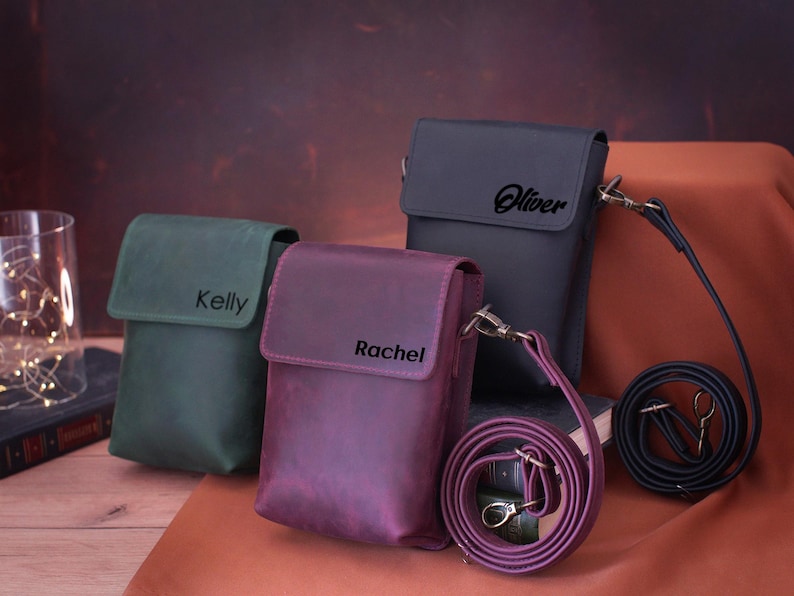 May include: Three leather crossbody bags in green, purple, and dark gray. Each bag has a flap closure and a name printed on the front: "Kelly", "Rachel", and "Oliver". The bags have adjustable straps and metal hardware.