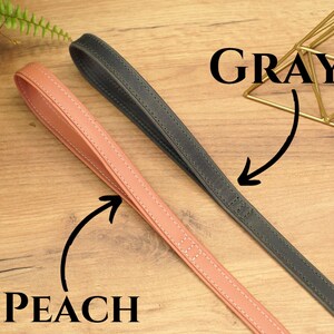 Puede incluir: Dos correas de cuero para perros, una de color melocot&oacute;n y otra gris, colocadas sobre una superficie de madera. Las correas est&aacute;n cosidas y tienen una textura lisa.