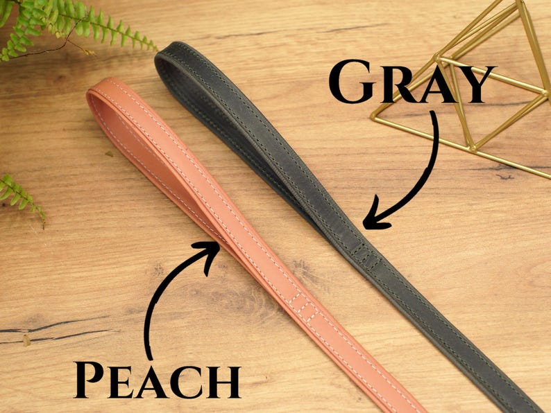 Puede incluir: Dos correas de cuero para perros, una de color melocot&oacute;n y otra gris, colocadas sobre una superficie de madera. Las correas est&aacute;n cosidas y tienen una textura lisa.