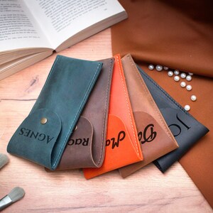 May include: Five leather pencil cases in different colors, each with a personalized name engraved on the front. The cases are arranged in a row, with the blue case at the front and the black case at the back. The names on the cases are Agnes, Rad, Ma, Dee, and Jo.