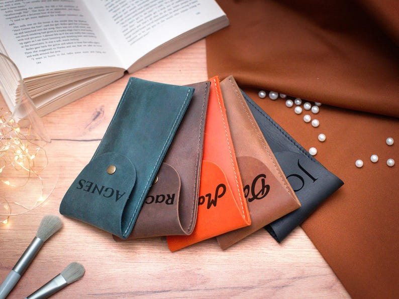 May include: Five leather pencil cases in different colors, each with a personalized name engraved on the front. The cases are arranged in a row, with the blue case at the front and the black case at the back. The names on the cases are Agnes, Rad, Ma, Dee, and Jo.
