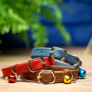 May include: Three cat collars with bells. The collars are made of leather and come in red, brown, and gray. The bells are gold, red, and blue.
