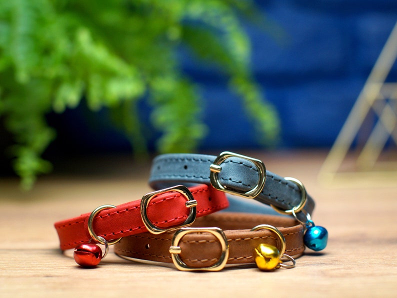 May include: Three cat collars with bells. The collars are made of leather and come in red, brown, and gray. The bells are gold, red, and blue.