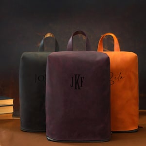 May include: Three personalized leather bags in black, burgundy, and orange. Each bag has a handle and a zipper at the bottom. The bags are embossed with initials or a name. The background is a dark brown.