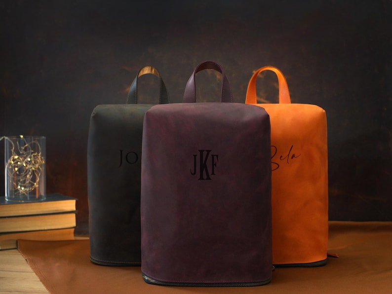 May include: Three personalized leather bags in black, burgundy, and orange. Each bag has a handle and a zipper at the bottom. The bags are embossed with initials or a name. The background is a dark brown.