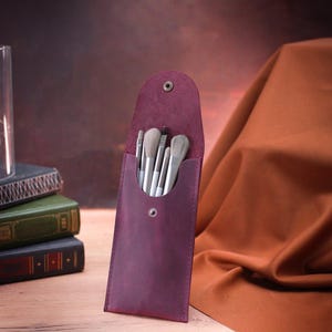 May include: A burgundy leather makeup brush holder with a snap closure. The holder is filled with a set of white makeup brushes.