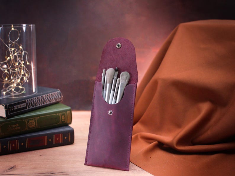 May include: A burgundy leather makeup brush holder with a snap closure. The holder is filled with a set of white makeup brushes.