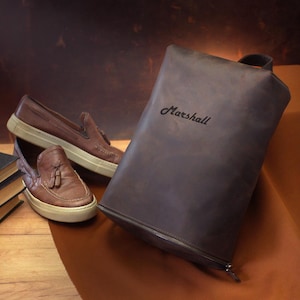 May include: A brown leather shoe bag with the name "Marshall" in script, a pair of brown leather loafers with tassels, and a stack of books. The shoes have white soles. The bag has a zipper.
