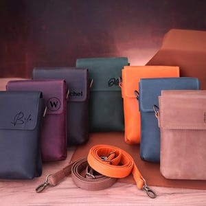 May include: A collection of leather crossbody bags in various colors, including navy, purple, green, orange, blue, and brown. Each bag features a flap closure and a shoulder strap. Some bags have personalized initials.