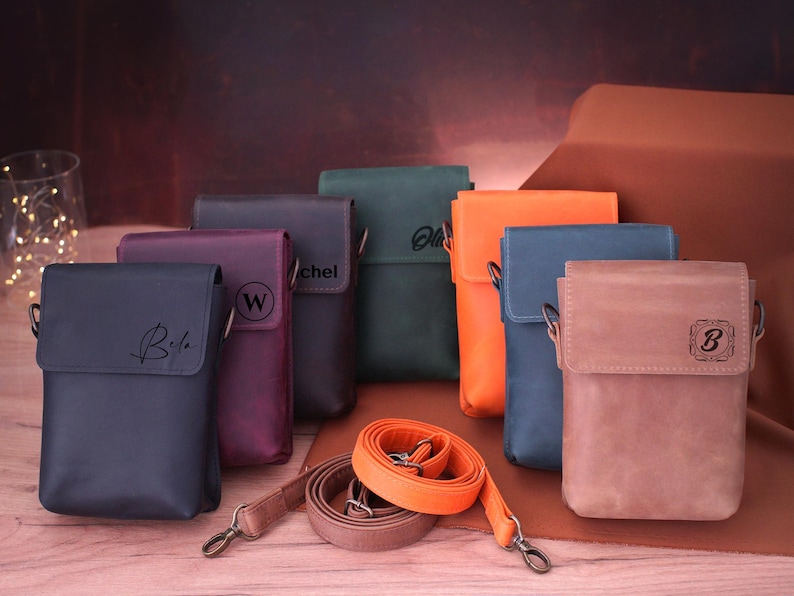 May include: A collection of leather crossbody bags in various colors, including navy, purple, green, orange, blue, and brown. Each bag features a flap closure and a shoulder strap. Some bags have personalized initials.
