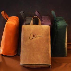 May include: A collection of leather tote bags in various colors, including orange, brown, green, and purple. The bags have a rectangular shape, a top handle, and a zipper closure. The word "Oliver" is written on the front of one bag.